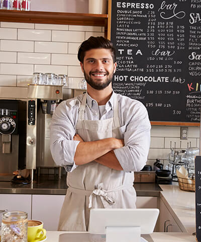 Ventajas para Autónomos - El dueño de una cafetería con los brazo cruzados sonriendo tras la barra de su negocio Ventajas para Autónomos - El dueño de una cafetería con los brazo cruzados sonriendo tras la barra de su negocio