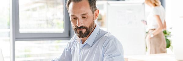 Programa Nómina - Manager de una empresa de mediana edad con barba y camisa azul trabajando en su portátil en la oficina